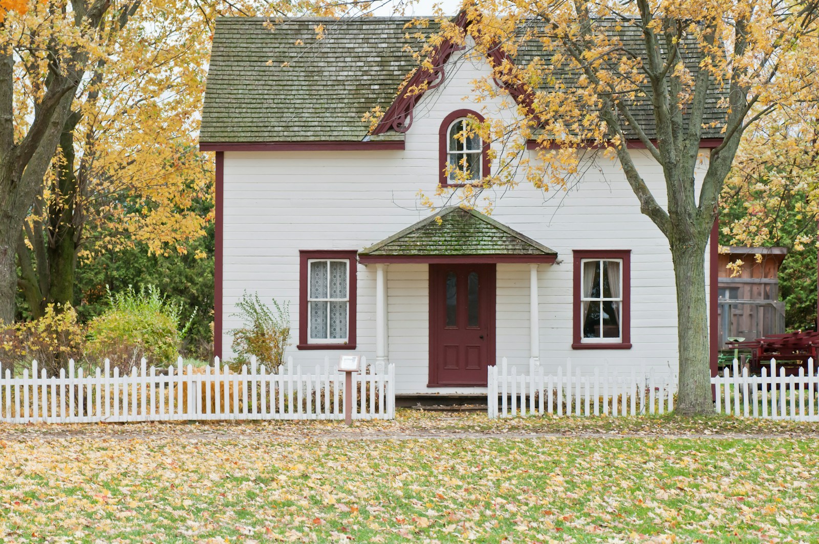 A white house with red trim behind a picket fence, framed by yellow maples and fallen leaves on the lawn